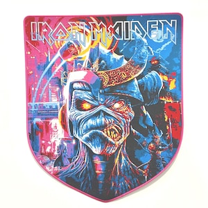 Puede incluir: Parche del logotipo de la banda Iron Maiden con una imagen azul y roja de Eddie, la mascota de la banda, en un entorno futurista. El parche tiene forma de escudo y tiene un borde rosa.