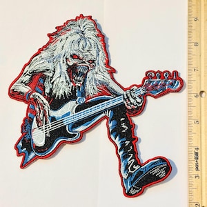 May include: An embroidered patch featuring a long-haired, sharp-toothed creature playing a black and blue bass guitar. The patch has a red border and is positioned next to a wooden ruler showing a height of approximately 16.5 cm.