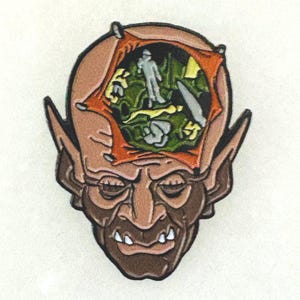 May include: A detailed enamel pin featuring a grotesque face with large ears and a sinister grin. The head is split open, revealing a green and yellow interior with a small figure. The pin is brown, orange, and green.