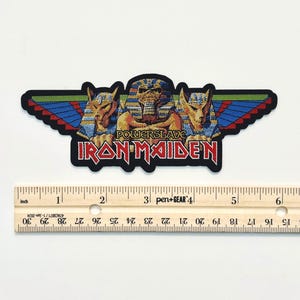 Puede incluir: Un parche bordado de Iron Maiden Powerslave que presenta una figura central de faraón flanqueada por dos cabezas de chacal con alas azules, verdes y rojas. El parche mide aproximadamente 14 cm de ancho, como muestra la regla debajo.