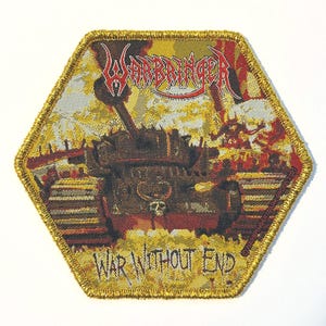 以下が含まれることがあります： 金色の縁取りが付いた六角形のパッチで、戦車のイラストが描かれています。「WARBRINGER」の文字が赤色で、「WAR WITHOUT END」の文字が白色で書かれています。背景は黄色、赤色、茶色の混合です。