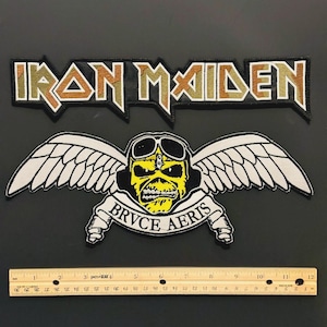 Könnte beinhalten: Zwei aufgestickte Aufnäher mit der Band Iron Maiden. Der obere Aufnäher trägt "IRON MAIDEN" in Gold und Olivgrün. Der untere Aufnäher zeigt einen gelben Schädel mit Fliegerbrille und weißen Flügeln, mit "BRVCE AERIS" auf einem Banner. Ein Lineal zeigt Messungen in cm.