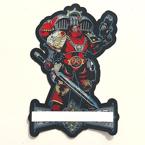 Pode incluir: Um emblema recortado apresentando um guerreiro com armadura vermelha em um traje futurista, segurando uma espada longa e uma arma de fogo. O guerreiro tem um emblema de caveira alada no ombro, uma fivela de cinto dourada e uma base decorativa.