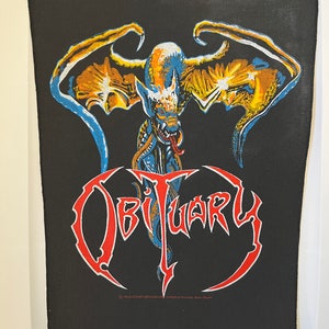 Könnte beinhalten: Schwarzer Bügel-Patch mit einer roten, blauen und gelben Grafik eines geflügelten Dämons mit dem Wort "Obituary" in roten Buchstaben.