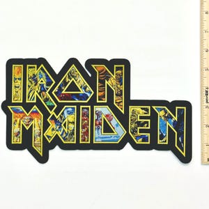 May include: A fabric patch featuring the Iron Maiden logo in a bold, block-letter style with a yellow border. The letters are filled with colorful, detailed illustrations of various characters. A wooden ruler shows the patch is 11 inches wide.