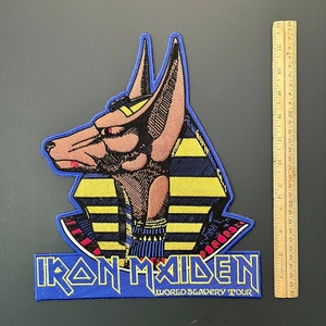 Puede incluir: Parche de la gira World Slavery Tour de Iron Maiden con una imagen bordada en azul y amarillo de una cabeza de chacal Anubis con el nombre de la banda y el título de la gira.