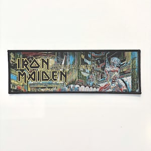 Puede incluir: Un parche rectangular bordado que presenta el logotipo de Iron Maiden y el texto Somewhere in Time. El diseño muestra un paisaje urbano futurista con una figura cíborg en tonos rojos y azules, todo dentro de un borde negro cosido.