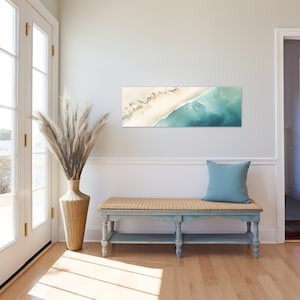 May include: A light blue bench with a woven straw seat and a blue pillow on top. The bench is in front of a white wall with a horizontal painting of an aerial view of a beach and ocean.