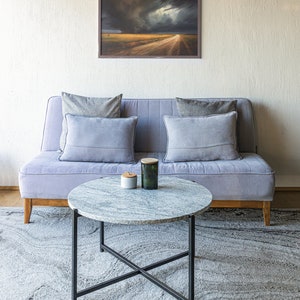 May include: A light gray sofa with gray pillows sits in front of a white wall with a framed landscape painting above it. A round marble coffee table with a black metal frame sits in front of the sofa on a gray rug.
