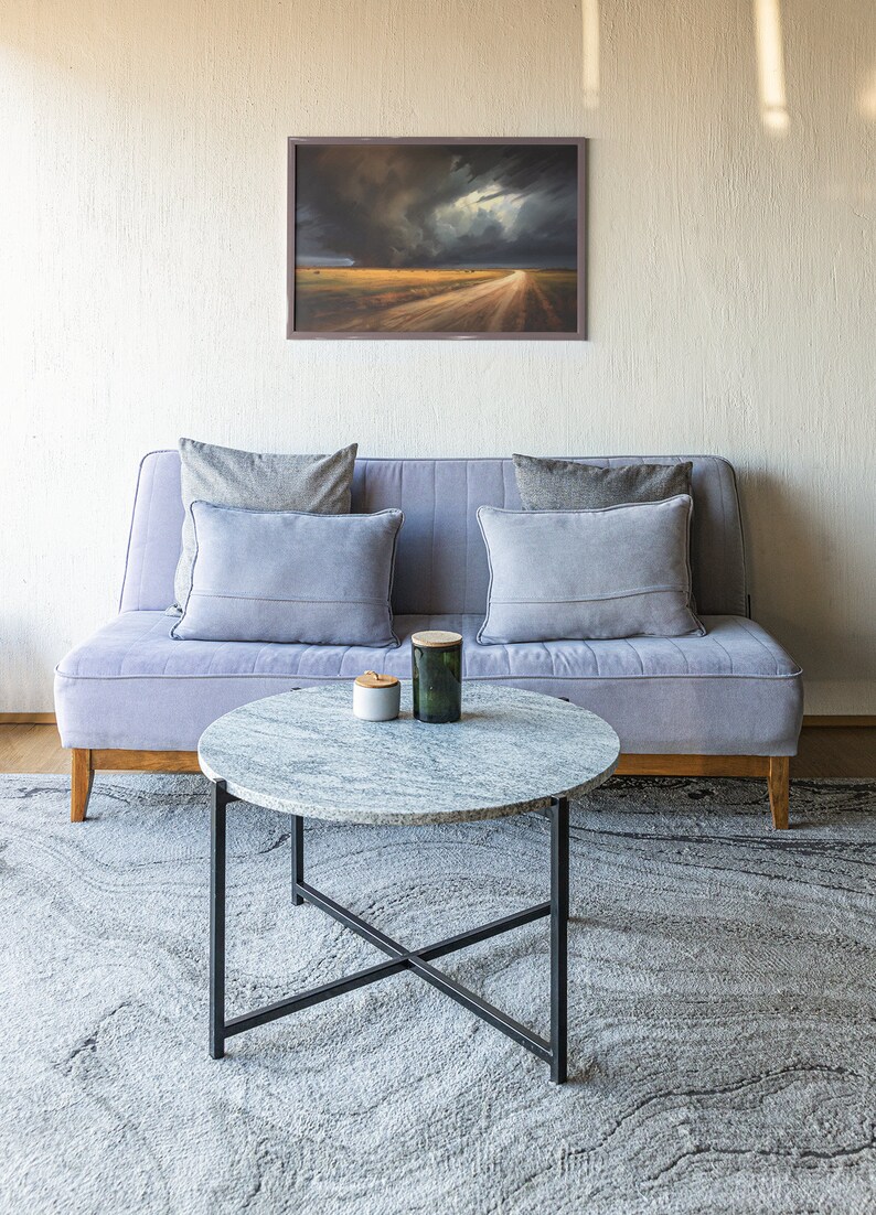 May include: A light gray sofa with gray pillows sits in front of a white wall with a framed landscape painting above it. A round marble coffee table with a black metal frame sits in front of the sofa on a gray rug.