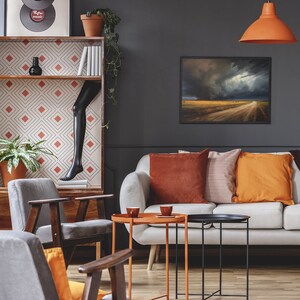 May include: A living room with a white sofa, two gray armchairs, and a black and white patterned rug. The sofa has orange and brown throw pillows. There are two coffee tables, one orange and one black. A framed picture of a stormy landscape hangs on the wall. A hanging lamp with an orange shade hangs from the ceiling. A bookshelf with a geometric pattern and a black mannequin leg is in the background.