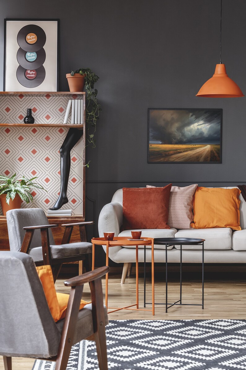 May include: A living room with a white sofa, two gray armchairs, and a black and white patterned rug. The sofa has orange and brown throw pillows. There are two coffee tables, one orange and one black. A framed picture of a stormy landscape hangs on the wall. A hanging lamp with an orange shade hangs from the ceiling. A bookshelf with a geometric pattern and a black mannequin leg is in the background.