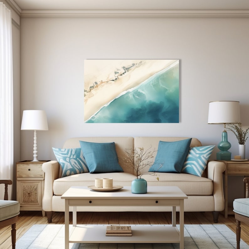 May include: A watercolor painting of a beach scene with a sandy shore and turquoise ocean water. The painting is framed and hanging on a wall above a beige sofa with blue pillows.