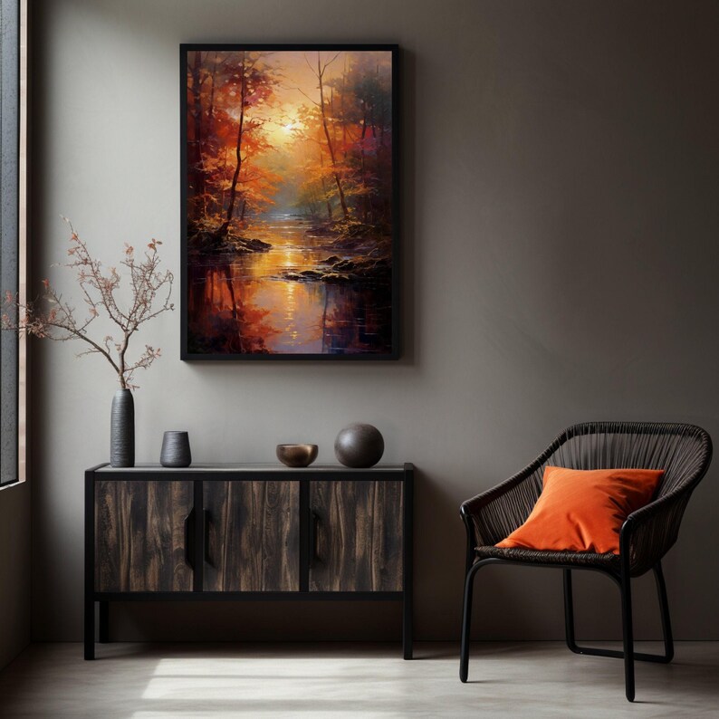 May include: A rectangular framed wall art print depicting a forest landscape with a stream at sunset, featuring warm orange, yellow, and deep brown tones. The artwork is displayed above a dark wood cabinet with a vase, two bowls, and a chair with an orange pillow.