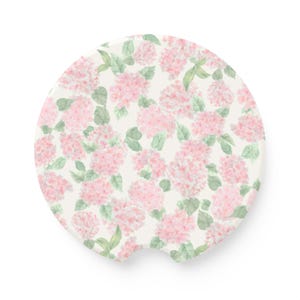 May include: A circular car coaster featuring a watercolor-style pattern of soft pink hydrangea flower clusters and light green leaves set against an off-white background, with a small semi-circular notch cut out from the bottom edge.