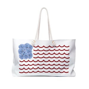 May include: A white tote bag with two rope handles featuring a graphic design of a blue hydrangea flower cluster in the upper left corner and horizontal rows of red wavy lines extending across the remainder of the bag's front surface.