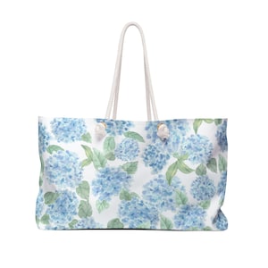 Könnte beinhalten: Eine rechteckige Tragetasche mit einem Aquarell-Druck von blauen Hortensienblüten und grünen Blättern auf weißem Grund. Die Tasche verfügt über zwei weiße Seilgriffe, die mit silberfarbenen Metallösen an der Oberkante befestigt sind.