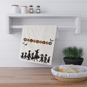 May include: A cream-colored kitchen towel featuring the word HALLOWEEN in alternating orange and black circles. Below the text is a black silhouette graphic of a group of children in costumes and a witch walking with pumpkin buckets and bats.