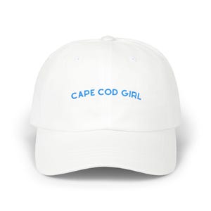 May include: A white baseball cap featuring the text CAPE COD GIRL embroidered in light blue thread across the front panel. The hat has a structured crown, a curved brim, and a button at the top centre, providing a clean and simple design.