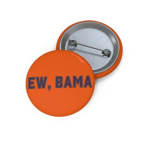 May include: A round orange pin-back button featuring the dark blue text EW, BAMA printed in a bold sans-serif font. The back of the button is silver-colored metal with a horizontal pin mechanism attached for securing the item to fabric or clothing.
