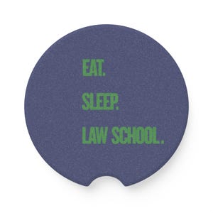 May include: A circular dark blue car coaster with a textured surface and a small semi-circular notch at the bottom edge. The text EAT. SLEEP. LAW SCHOOL. is printed in a bold, bright green sans-serif font across the center of the coaster.