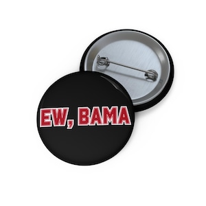 May include: A round pin-back button with a black background and the text EW, BAMA in red capital letters with a white outline. The back of the button is silver-colored metal with a horizontal pin mechanism for attachment to clothing or fabric.