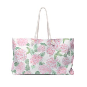 Könnte beinhalten: Eine große Tragetasche mit einem Aquarell-Druck von rosa Hortensienblüten und grünen Blättern auf weißem Grund. Die Tasche verfügt über zwei weiße Seilgriffe, die mit goldfarbenen Metallbeschlägen an der Oberseite des rechteckigen Körpers befestigt sind.