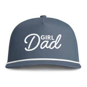 May include: A slate blue baseball cap featuring the white embroidered text "GIRL Dad" across the front panel. The cap includes a white braided cord trim running horizontally across the base of the crown, just above the curved brim.