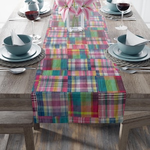 May include: A rectangular table runner featuring a patchwork design of multicolored plaid fabric squares in shades of pink, teal, yellow, and blue, centered on a wooden dining table with place settings and a glass vase of pink lilies.