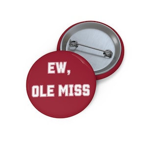 Pode incluir: Um botton redondo com fundo vermelho escuro e texto branco que diz EW, OLE MISS. O botton apresenta uma parte traseira de metal prateado com um mecanismo de alfinete horizontal para fixação em roupas ou acessórios de tecido.