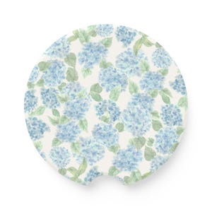 May include: A circular car coaster featuring a watercolor pattern of light blue hydrangea flower clusters and soft green leaves on a white background. The coaster has a small semi-circular notch cut out from the bottom edge for easy removal.