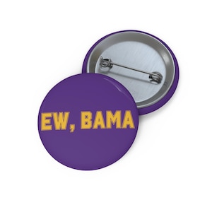 May include: A round pinback button featuring a solid purple background with the text EW, BAMA printed in bold yellow capital letters. The back of the button is metallic silver and includes a horizontal pin mechanism for fastening to clothing.