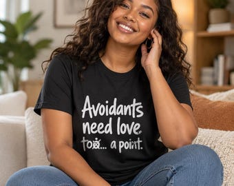 Può includere: Una t-shirt nera a maniche corte con testo bianco che recita: Avoidants need love too... a point. Il design include una piccola grafica di un guscio di tartaruga integrata nel testo, che sostituisce specificamente la lettera o.
