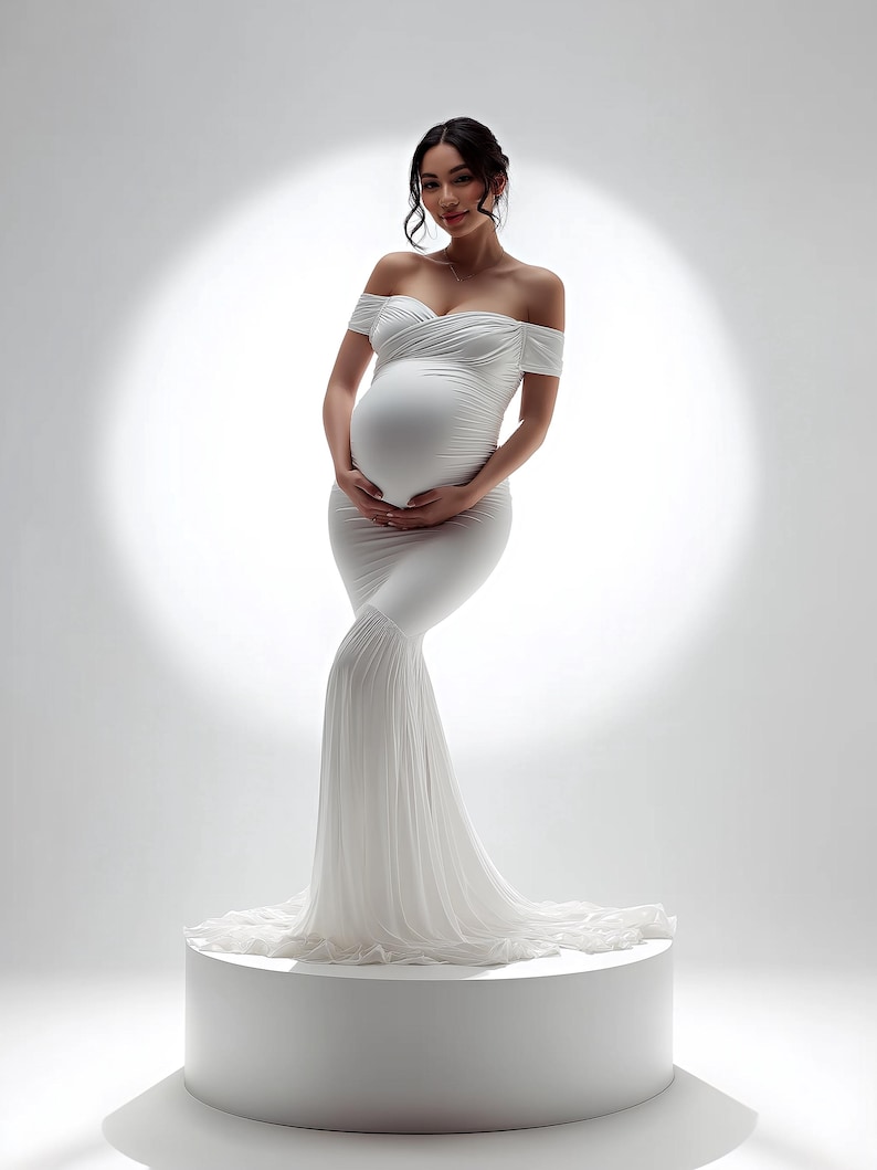 May include: A white off-the-shoulder maternity gown with a mermaid silhouette. The dress features a fitted bodice, a flowing skirt, and a train. The model is standing on a white pedestal in front of a bright white background.