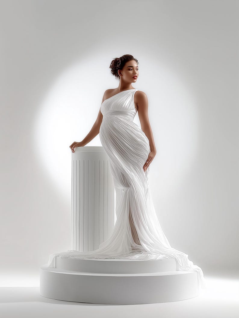 May include: A white, one-shoulder gown with a flowing, pleated design. The dress features a high slit and a long train. The model is standing on a white pedestal next to a white column. The overall theme is maternity fashion.