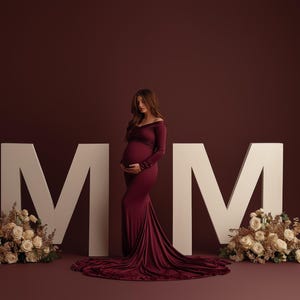 May include: A pregnant person in a burgundy off-the-shoulder gown stands before large white letters spelling "MM". Floral arrangements flank the letters. The background is a solid burgundy color.