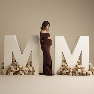 May include: A pregnant person in a brown off-the-shoulder dress stands between two large white letter "M" structures. The letters are decorated with beige and brown flowers, creating a soft, elegant backdrop.
