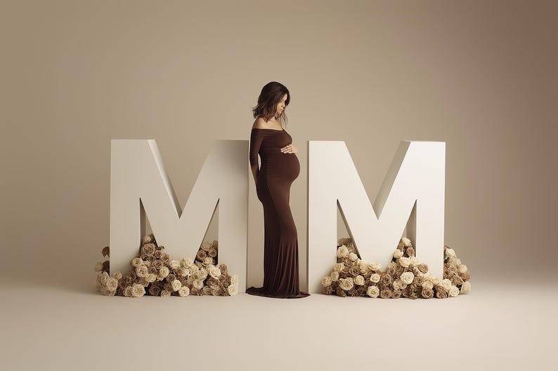 May include: A pregnant person in a brown off-the-shoulder dress stands between two large white letter "M" structures. The letters are decorated with beige and brown flowers, creating a soft, elegant backdrop.