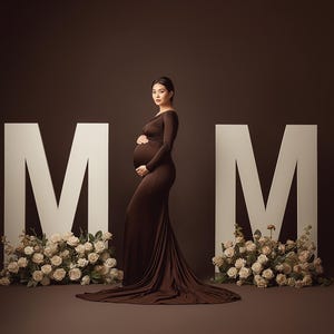 May include: A pregnant person in a long, brown gown poses between two large white "M" letters. Cream-colored roses adorn the base of each letter. The background is a solid, dark brown.