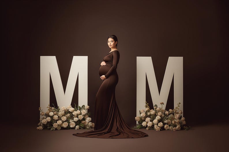 May include: A pregnant person in a long, brown gown poses between two large white "M" letters. Cream-colored roses adorn the base of each letter. The background is a solid, dark brown.