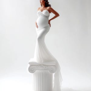 May include: A white, strapless maternity gown with a flowing train. The dress features a fitted bodice with delicate embellishments and a ruched skirt. The model is standing on a white column, posing against a white backdrop.
