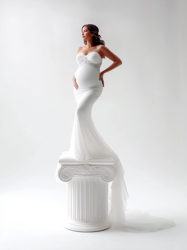 May include: A white, strapless maternity gown with a flowing train. The dress features a fitted bodice with delicate embellishments and a ruched skirt. The model is standing on a white column, posing against a white backdrop.