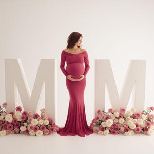 May include: A woman in a long, burgundy maternity gown stands between large white letters spelling "MOM." Pink and white roses adorn the base of the letters. The off-the-shoulder dress highlights the woman's pregnancy.