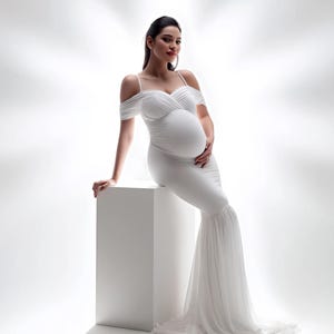 May include: A white maternity gown with off-the-shoulder sleeves and a flowing, layered skirt. The dress is form-fitting through the torso, accentuating the pregnant figure. The gown is worn by a person sitting on a white cube.