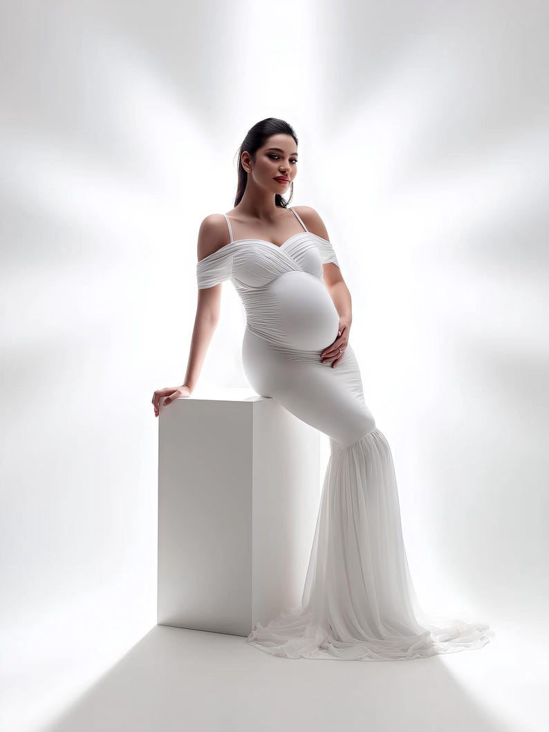 May include: A white maternity gown with off-the-shoulder sleeves and a flowing, layered skirt. The dress is form-fitting through the torso, accentuating the pregnant figure. The gown is worn by a person sitting on a white cube.