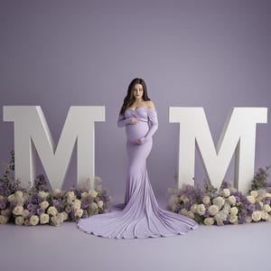 May include: A woman in a lavender off-the-shoulder maternity gown stands between two large white "M" letters. The letters are flanked by floral arrangements of white and purple flowers. The background is a solid lavender color.