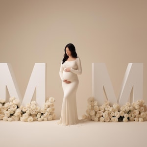 May include: A pregnant person in a cream-colored gown stands between large white letters spelling "MOM". The letters are decorated with cream-colored roses, creating a soft, elegant maternity photo setting.
