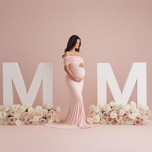May include: A pregnant person in a light pink off-the-shoulder gown stands between two large white "M" letters. The letters are decorated with white and pink roses, set against a soft pink backdrop.
