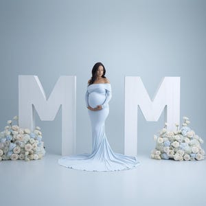 May include: A woman in a light blue off-the-shoulder maternity gown stands between two large white letter "M" props. Floral arrangements in pastel colors are at the base of each letter. The gown has a long train.