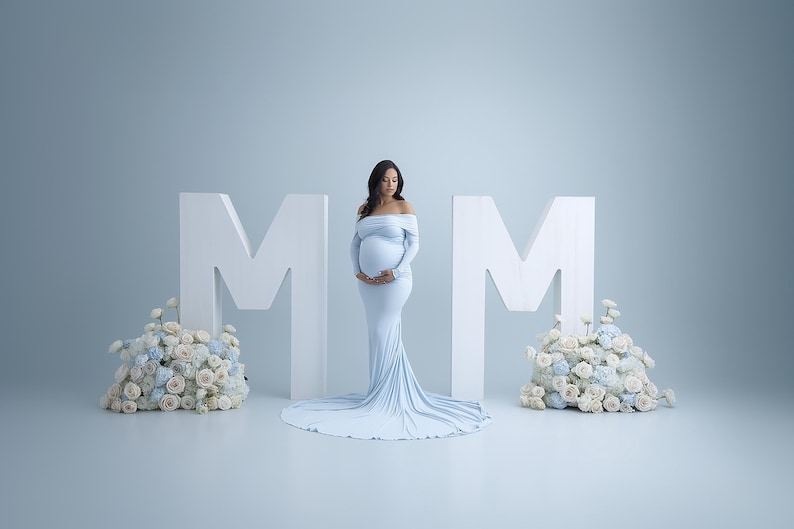 May include: A woman in a light blue off-the-shoulder maternity gown stands between two large white letter "M" props. Floral arrangements in pastel colors are at the base of each letter. The gown has a long train.